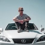 A trendy man sits confidently on a white Lexus, exuding style in a sunlit outdoor setting.